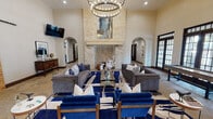 Located in Grapevine, Texas, this single-story space showcases a refined Hill Country-inspired architectural design that seamlessly blends rustic charm with modern elegance. The centerpiece of the main living area is a dramatic floor-to-ceiling limestone fireplace, flanked by arched doorways and anchored by a striking circular chandelier that illuminates the soaring ceiling with exposed dark wood beams. Natural stone tile flooring flows throughout the space, complementing the warm, neutral palette accented by rich blue furnishings. Large multi-pane French doors and windows line the walls, inviting abundant natural light and offering views of the outdoors. The spacious living area is thoughtfully arranged with tufted gray sofas and bold blue accent chairs around a glass-top coffee table, creating an inviting and sophisticated gathering environment. A gourmet kitchen features rich dark wood cabinetry, a professional-grade stainless range, a substantial stone hood surround, and a generously sized island topped with deep brown marble-finished stone, complete with blue velvet bar seating. Arched passageways connect the various areas of the space, reinforcing the cohesive architectural language throughout. The overall ambiance is warm, grand, and welcoming, combining high-end finishes with thoughtful design details at every turn.
