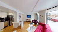 This single-floor unit combines contemporary design with bold, eclectic styling across its thoughtfully arranged layout. The open-concept living area serves as the heart of the space, featuring warm hardwood flooring, crisp white walls, and rail track lighting that runs across the ceiling. A large floor-to-ceiling window at one end of the living room draws in natural light and provides an outdoor view, while a wall-mounted flat-screen TV and a gold-accented console anchor the space. The seating area is furnished with a striking red sofa and a purple accent chair, adding vivid pops of color against the neutral backdrop.

The kitchen is defined by white flat-front cabinetry, dark granite countertops, and a bold black subway tile backsplash, creating a sharp contrast. A partial white partition wall separates the kitchen from the living area, providing both visual delineation and a convenient breakfast bar surface.

The bedroom continues the hardwood flooring and features a ceiling fan with integrated track lighting, a wall-mounted TV, and ample space for a full dresser and nightstands.

The bathroom is finished entirely in white, with wood-look flooring, a single-sink vanity, a tub-shower combination with white tile surround, and artful decorative accents that give it a gallery-like quality.