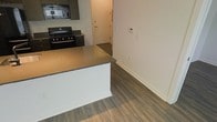 This single-floor space showcases a thoughtfully designed open-concept layout that seamlessly connects the kitchen and living areas. The kitchen is equipped with dark cabinetry, a black gas range with an overhead microwave, and a peninsula-style island featuring a dark stone countertop with a crisp white base, providing both additional prep space and a natural visual divider between the kitchen and the adjoining living area. The consistent dark hardwood-style flooring runs throughout the space, adding warmth and continuity to the design. Neutral white walls keep the interior feeling bright and clean, complementing the modern aesthetic established by the dark fixtures and cabinetry. A thermostat panel is mounted on the wall, indicating a climate-controlled environment. The entryway, visible through the kitchen area, features a solid door and offers a straightforward, functional transition into the unit. The overall ambiance is contemporary and minimalist, with a cohesive palette of dark and light tones that creates a sleek, urban feel suited to modern living.