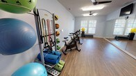 Located at 205 McCaskill Rd in Pinehurst, NC, this single-floor fitness studio is purpose-built to support a wide range of exercise modalities. The space features durable wood-style flooring throughout, providing a clean and resilient surface suited for both cardio and strength training activities.

The studio is outfitted with an organized equipment rack holding an array of resistance bands, medicine balls in varying weights, foam rollers, and stability balls in multiple sizes, ensuring versatility for individual and group workouts alike. Multiple spin bikes are positioned to allow for cycling classes or independent cardio sessions.

A prominent floor-to-ceiling mirror wall spans one side of the room, enabling users to monitor their form during exercise and giving the space a sense of openness and depth. Portable folding mats on wheeled frames offer flexibility for floor-based workouts and can be easily repositioned or stored.

Natural light enters through several windows fitted with white blinds, contributing to a bright and energizing atmosphere. Ceiling fans with overhead lighting fixtures maintain air circulation and comfort throughout the space. The light blue-gray painted walls create a calm, focused environment conducive to physical activity.

The open floor plan allows for ample movement space, making the studio well-suited for group fitness classes, personal training sessions, or individual exercise routines.