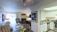 This single-story residence showcases an open-concept floor plan that seamlessly integrates the living, dining, and kitchen areas into one cohesive and functional space. The living room serves as a central focal point, anchored by a fireplace framed by a dark wood media console and complemented by contemporary abstract wall art. Neutral gray walls provide a versatile backdrop, while hardwood-style flooring adds warmth and continuity throughout the space. Pendant lighting and a ceiling fan with integrated lighting fixtures illuminate the open layout, enhancing both form and function. The dining area transitions naturally from the living space, offering a casual setting ideal for everyday meals or entertaining. Adjacent to the dining zone, the kitchen features white cabinetry, a double-basin sink, and light countertops that create a clean, bright workspace. A half-wall partition subtly defines the kitchen without closing it off from the rest of the space. The textured popcorn ceiling adds a distinctive finish overhead. Large windows dressed with light, airy curtains invite natural light into the living area, contributing to the overall welcoming ambiance of this thoughtfully designed residence.