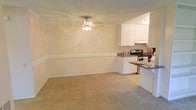 This single-floor space showcases an open-concept layout that seamlessly connects the living area to the kitchen. The living area is finished with wall-to-wall beige carpeting, creating a warm and neutral foundation throughout. A decorative ceiling light fixture serves as a subtle focal point overhead, adding a touch of elegance to the room. A chair rail molding runs along the walls, providing a classic architectural detail that adds visual structure to the space.

The kitchen is situated adjacent to the living area and features crisp white cabinetry that extends to the upper walls, offering ample storage. Dark countertops provide a contrasting accent against the white cabinets, while a stainless steel electric range serves as the primary cooking appliance. A peninsula or breakfast bar countertop borders the kitchen, creating a natural division between the two areas while maintaining the open feel of the layout. The transition from carpet to smooth flooring delineates the kitchen zone clearly. Overall, the space presents a clean, functional, and cohesive interior with a light, neutral color palette throughout.