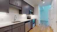 This contemporary single-floor residence showcases a thoughtfully designed layout with clean lines and modern finishes throughout. The kitchen is a standout feature, equipped with stainless steel appliances including a refrigerator, electric range, built-in microwave, and dishwasher. Sleek dark gray cabinetry with brushed metal hardware extends from floor to ceiling, complemented by white quartz countertops and a classic subway tile backsplash with under-cabinet lighting. The single-basin sink with a modern pull-down faucet adds a functional touch.

Light-toned hardwood flooring runs seamlessly throughout the living areas, creating a warm and cohesive aesthetic. The main living area is particularly impressive, featuring soaring ceilings and a wall of large windows that flood the space with natural light and provide views of the surrounding streetscape.

The bathroom is finished in a clean, minimal style with light tile flooring, a full-size soaking tub with an overhead shower, and a gray vanity with a white countertop. The entry foyer opens directly to the kitchen and living areas, offering a practical and open flow. Overall, the space combines modern urban design with functional living essentials in a bright, well-finished interior.