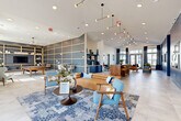 Located at 191 Commerce Dr in Villa Rica, Georgia, this thoughtfully designed community clubhouse offers a generous single-floor layout that balances social gathering spaces with functional amenities. The interior makes a strong first impression with its soaring vaulted ceilings, large-format tile flooring, and a cohesive navy blue and warm wood color palette that flows throughout the space.

The main lounge area serves as the heart of the clubhouse, furnished with a tan leather sofa, mid-century style accent chairs in blue patterned upholstery, and a bold patterned area rug. Statement sculptural pendant lighting hangs overhead, while floor-to-ceiling windows dressed in two-tone navy drapes flood the space with abundant natural light and views of the surrounding exterior.

Adjacent to the lounge is a versatile game and recreation room, featuring a pool table, open shelving with decorative displays, and casual seating arrangements set against a textured blue accent wall with gold-trimmed grid paneling. A substantial live-edge wood communal bar table with leather and brass barstools provides an ideal co-working or gathering spot nearby.

A dedicated beverage station with navy cabinetry, hexagonal tile backsplash, a beverage refrigerator, and a Keurig coffee setup adds everyday convenience. Private offices with frosted-glass doors and a workstation with outdoor views offer quiet, functional workspace. Curated music-themed artwork adorns the walls, adding cultural character throughout.