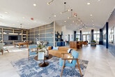 Located at 191 Commerce Dr in Villa Rica, Georgia, this thoughtfully designed community clubhouse offers a generous single-floor layout that balances social gathering spaces with functional amenities. The interior makes a strong first impression with its soaring vaulted ceilings, large-format tile flooring, and a cohesive navy blue and warm wood color palette that flows throughout the space.

The main lounge area serves as the heart of the clubhouse, furnished with a tan leather sofa, mid-century style accent chairs in blue patterned upholstery, and a bold patterned area rug. Statement sculptural pendant lighting hangs overhead, while floor-to-ceiling windows dressed in two-tone navy drapes flood the space with abundant natural light and views of the surrounding exterior.

Adjacent to the lounge is a versatile game and recreation room, featuring a pool table, open shelving with decorative displays, and casual seating arrangements set against a textured blue accent wall with gold-trimmed grid paneling. A substantial live-edge wood communal bar table with leather and brass barstools provides an ideal co-working or gathering spot nearby.

A dedicated beverage station with navy cabinetry, hexagonal tile backsplash, a beverage refrigerator, and a Keurig coffee setup adds everyday convenience. Private offices with frosted-glass doors and a workstation with outdoor views offer quiet, functional workspace. Curated music-themed artwork adorns the walls, adding cultural character throughout.