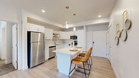This well-appointed 2-bedroom, 2-bathroom residence offers a thoughtfully designed single-level layout with modern finishes throughout. The open-concept kitchen and living area serves as the heart of the home, featuring crisp white cabinetry, stainless steel appliances, a built-in microwave, and a central island with bar seating illuminated by stylish pendant lighting. Light wood-style flooring flows seamlessly through the main living areas, enhancing the bright, airy atmosphere. The living room opens directly to a private balcony through sliding glass doors with vertical blinds, allowing for abundant natural light. The primary bedroom offers a comfortable, carpeted retreat with a dedicated closet, and connects to a full en-suite bathroom appointed with a wide vanity, ample cabinetry, and recessed lighting. The second bedroom is equally well-sized, featuring large windows and plush carpeting. A second full bathroom serves the remaining rooms. A convenient in-unit laundry area with washer and dryer and overhead shelving adds practical functionality. A hallway and dedicated closet provide additional storage. The overall design palette of neutral tones, recessed lighting, and clean architectural lines creates a cohesive, contemporary living environment.