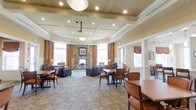 This refined single-floor communal space offers a welcoming blend of lounge and dining areas, thoughtfully designed to foster social interaction and relaxation. The room's most striking architectural feature is its elegant coffered ceiling, highlighted by recessed lighting and a central pendant fixture that bathes the space in a warm, inviting glow. A ceiling-mounted projector adds a functional touch for events and presentations.

At the heart of the room stands a handsome stone fireplace, framed by soft yellow walls and flanked by large windows dressed in rich amber-toned drapes. These oversized windows flood the interior with natural light, creating an airy and uplifting atmosphere throughout the day.

The space is furnished with a thoughtful mix of plush lounge seating and traditional dining tables and chairs, accommodating both casual conversation and more formal gatherings. A patterned area rug anchors the central seating area, adding texture and warmth underfoot.

Architectural details such as white wainscoting, decorative columns, and arched doorways lend the space a classic, sophisticated character. Potted plants and table lamps are strategically placed to enhance the homey ambiance. Overall, this space is well-appointed and designed to serve as a vibrant hub for community life.