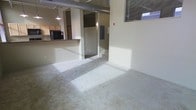This single-floor space showcases an industrial-influenced, open-concept design that blends functional living areas with loft-style architectural details. Exposed cylindrical ductwork runs along the ceiling, adding a distinctive industrial character that contrasts with the neutral, light-toned walls throughout. The flooring in the main living area consists of plush, light-colored carpet, contributing to a warm and comfortable atmosphere.

A peninsula-style kitchen counter separates the cooking area from the main open space, offering a casual bar-style setup. The kitchen features light wood cabinetry and a dark refrigerator visible in the background, complemented by a hanging pendant light that adds focused illumination. The elevated counter provides a natural division between spaces without fully closing off the open-plan layout.

A partially visible corridor or secondary room leads further into the unit, lit by an additional pendant light, suggesting defined zones within the open layout. Windows fitted with horizontal blinds allow natural light to filter into the space, brightening the neutral interior. A fire safety device is mounted on the wall, and the overall aesthetic leans toward a utilitarian yet livable urban design.