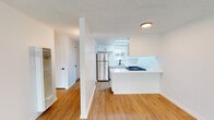 This single-floor unit offers a clean, modern aesthetic defined by bright white walls, light hardwood-style flooring, and a textured ceiling throughout the main living areas. A wide entryway hallway with a wall sconce leads into the open-concept living and kitchen space, where natural light filters in through slatted blinds on a large front-facing window. The living area is spacious and open, providing a versatile canvas for various furniture arrangements.

The kitchen is thoughtfully designed with crisp white cabinetry featuring black hardware, gleaming white countertops, and a full suite of stainless steel appliances including a refrigerator, gas range, and over-the-range microwave. A peninsula counter separates the kitchen from the living area, enhancing the open flow of the space. A skylight-style ceiling fixture provides additional illumination in the kitchen, and a window above the sink brings in natural light.

The bedroom features soft neutral carpeting and clean white walls, offering a quiet and comfortable retreat. The bathroom is finished with white fixtures throughout, including a full tub-shower combination, a vanity with storage, and a small frosted window for natural ventilation. A large wall-mounted HVAC unit services the living area, ensuring year-round climate control. Overall, the space presents a fresh, move-in ready environment.