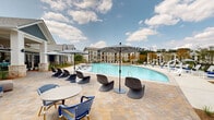 Located at 300 Highway 81 in McDonough, Georgia, this resort-style outdoor amenity space is designed to deliver an upscale leisure experience. At its centerpiece is a generously sized swimming pool with a curvilinear shape, surrounded by a wide, paver-stone deck lined with lounge chairs and shade umbrellas in a classic navy-and-white stripe pattern. Carefully curated landscaping — including ornamental shrubs, native grasses, and mulched garden beds — frames the pool perimeter, adding greenery and visual texture throughout the space.

Adjacent to the pool, a spacious covered pavilion serves as the social hub of the area. Featuring a coffered ceiling with ceiling fans, the pavilion is open on multiple sides to allow natural light and views of the surrounding landscape. Inside, a central bar area finished with patterned tile and a sleek countertop is complemented by cobalt blue barstools and pendant lighting. Comfortable seating arrangements — including woven sofas with blue and gray accent pillows alongside dining tables with black chairs — provide ample space for both relaxation and gathering.

An open-air covered terrace extends the pavilion, offering additional café-style seating with views toward lush green grounds and the broader community. Black traditional-style lamp posts punctuate the walkways, adding a charming architectural detail throughout this well-appointed outdoor amenity destination.