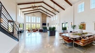 Located at 4130 High Mountain Road NE in Huntsville, Alabama, this striking single-floor space offers a generously sized open interior defined by dramatic vaulted ceilings with exposed dark wood beams. The architectural design blends modern farmhouse and contemporary elements throughout. Polished concrete floors reflect abundant natural light streaming in through floor-to-ceiling glass window walls, providing sweeping views of the surrounding landscape and an adjacent outdoor terrace.

A towering stacked-stone fireplace serves as the central focal point of the main gathering area, rising to the full height of the vaulted ceiling with a mounted flat-screen television above. Distinct functional zones are thoughtfully arranged across the open floor plan, including multiple lounge seating areas with stylish upholstered and leather furnishings, a formal dining area with a long table and leather chairs, and a bar or refreshment area.

Large industrial-style planters filled with lush snake plants are strategically placed throughout to define spaces and add organic warmth. Statement chandeliers and modern wall sconces provide layered lighting. A light wood staircase with a sleek metal railing leads to an upper level. The overall ambiance is bright, airy, and sophisticated, making this a highly functional and visually impressive venue.