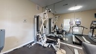 This compact yet fully equipped fitness center offers a comprehensive workout environment designed to support a range of exercise routines. The space features dark, durable flooring throughout, providing a practical and resilient surface suited for high-traffic use. Neutral-toned walls and white baseboards give the room a clean, functional aesthetic.

The strength training area is anchored by a multi-station cable machine, accompanied by a set of dumbbells for free weight exercises. Large wall mirrors line one side of the room, visually expanding the space and allowing users to monitor their form during workouts. A wall-mounted thermostat ensures climate comfort year-round.

The cardio section is well-appointed with a treadmill, an elliptical machine, and a recumbent bike, offering a variety of low- and high-impact exercise options. A wall-mounted flat-screen television is positioned for easy viewing from the cardio equipment, enhancing the workout experience.

Natural light filters into the space through a window and a glass-paned door fitted with horizontal blinds, offering views of surrounding greenery and providing a refreshing connection to the outdoors. The door also allows for convenient direct access to the exterior. Overall, this fitness center delivers a practical and inviting environment for daily exercise.