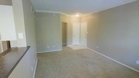 This single-floor space presents a well-maintained interior defined by its neutral beige walls and plush wall-to-wall carpeting, creating a warm and understated ambiance. Crown molding runs along the perimeter of the ceiling, adding a refined architectural detail that elevates the overall aesthetic. The ceiling height provides a comfortable sense of openness within the main living area. A flush-mount ceiling light fixture offers practical illumination, keeping the space bright and functional. White baseboards and door trim provide crisp contrast against the muted wall tones, contributing to a clean, cohesive look. Two white six-panel doors are visible — one appearing to serve as an entry or closet door, and another leading to an additional room or hallway beyond. The layout suggests a straightforward, functional flow between spaces. Electrical outlets are positioned along the walls at standard intervals, and a ventilation register is visible near the ceiling, indicating a central HVAC system. Overall, the space is move-in ready with a neutral palette that allows for versatile decorating possibilities.