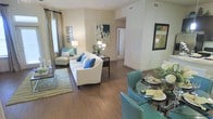 This single-story space showcases an open-concept layout that seamlessly integrates the living and dining areas into one cohesive, functional environment. The hardwood-style flooring flows continuously throughout, lending warmth and visual continuity to the space.

The living area is anchored by a white sofa adorned with teal and blue accent pillows, complemented by a cushioned armchair nearby. A patterned area rug defines the seating zone, while a table lamp and decorative wall art add character to the neutral, beige-toned walls. A window dressed with floor-length curtains allows natural light to filter into the room.

Adjacent to the living area, the dining space features a glass-top table surrounded by teal upholstered chairs, creating a bold yet cohesive color story that ties both zones together. Decorative place settings and fresh floral arrangements enhance the polished presentation.

A hallway visible in the background suggests additional rooms beyond, while ceiling vents indicate a central HVAC system. The overall ambiance is bright, modern, and welcoming, characterized by a clean neutral palette enlivened by consistent teal accents.