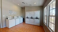 This single-floor communal laundry facility at 3245 E University Ave in Las Cruces, New Mexico, is designed for convenience and efficiency. The space features warm terracotta tile flooring throughout, providing a clean and durable surface suited to high-traffic use. The walls are finished in neutral taupe tones, complemented by a textured ceiling with recessed lighting that ensures the room is well-lit at all times.

The laundry room is outfitted with a row of top-loading washing machines along one wall, each equipped with individual controls and payment interfaces for easy operation. Opposite the washers, a bank of stacked commercial dryer units maximizes the use of vertical space, providing ample drying capacity. Instructional signage is posted near the machines to guide users through the washing process.

Large multi-pane windows along one wall allow generous natural light to filter into the space, offering views of the surrounding outdoor area. A separate entry corridor with glass-panel French doors connects the laundry room to an adjacent hallway, ensuring smooth circulation. A fire safety extinguisher is visibly mounted near the entrance, reflecting the facility's attention to safety standards. The overall layout is open and well-organized, making for a practical and welcoming shared laundry experience.