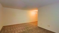 This single-floor space presents a clean, understated interior with a straightforward layout. The main living area features wall-to-wall carpet in a neutral tan tone, providing a warm and comfortable foundation underfoot. The walls are finished in a crisp off-white, contributing to a bright and open atmosphere throughout.

Transitioning from the main living area, a separate dining space is visible, distinguished by its warmer ambient lighting and a modest chandelier that serves as a subtle focal point. The shift from carpeted flooring to what appears to be a hard surface floor in the dining area adds a functional distinction between the two spaces.

The ceiling is smooth and standard in height, maintaining a consistent finish across both areas. Baseboards run along the perimeter of the rooms, adding a clean, finished edge to the walls. Electrical outlets are positioned at standard intervals along the walls, and the overall layout appears practical and efficient.

The neutral color palette and simple finishes throughout give this space a versatile, move-in-ready quality, offering a blank canvas well-suited to a variety of furnishing styles and personal touches.