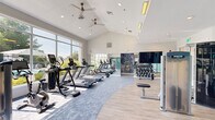 Located at 100 Hunsberger Dr in Limerick, PA, this fitness center offers a thoughtfully designed single-floor layout that accommodates a full range of workout options. The space is defined by its striking vaulted ceilings, which soar overhead and are complemented by dual ceiling fans, recessed lighting, and supplemental overhead heating elements — creating a bright, airy atmosphere year-round.

A defining architectural feature is the expansive floor-to-ceiling window wall that faces the outdoor pool area, flooding the cardio zone with abundant natural light and providing motivating views of the surrounding landscape. The flooring transitions purposefully between a soft blue patterned carpet — anchoring the cardio equipment area — and light wood-look hard flooring in the strength training zones, creating a clear visual distinction between functional areas.

The cardio section is equipped with treadmills, ellipticals, stationary bikes, and a rowing machine. The strength training area features a comprehensive dumbbell rack displayed beneath a large flat-screen TV, multiple selectorized cable and resistance machines, and additional free-weight benches. A mirrored wall with a bold tropical-print accent panel adds depth and style to this zone. A glass-paneled partition with cable rail detailing separates a smaller ancillary area, adding a modern architectural touch. The walls are finished in a calming aqua tone, enhancing the welcoming, energizing ambiance of the space.