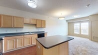Located at 100 2nd Ave in downtown Des Moines, Iowa, this one-bedroom, one-bathroom unit offers a practical and comfortable layout across a single floor. The kitchen is a standout feature, thoughtfully designed with an abundance of light wood cabinetry providing generous storage, dark countertops, and a full suite of black appliances including a refrigerator, range, microwave, and dishwasher. Rich hardwood-style flooring runs throughout the kitchen, and a peninsula counter extends the workspace while creating a natural boundary between the kitchen and the carpeted living area. The open layout of the main living space is airy and functional, with neutral tones throughout that create a clean, versatile canvas. The bedroom is a comfortable size, carpeted for warmth, and features a window with blinds that allows natural light to filter in. A dedicated closet provides additional storage. The bathroom is well-appointed with a wooden vanity with under-sink storage, a large mirror, wall lighting, and a neutral tile-style floor. The unit also includes a utility panel accessible from the bedroom area. Overall, the space presents a straightforward, efficient design well-suited for everyday living in a central Des Moines location.