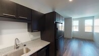 This single-floor unit offers a clean, contemporary design throughout, characterized by neutral gray walls, recessed lighting, and durable wood-look flooring that flows seamlessly from room to room. The kitchen is a standout feature, outfitted with rich dark espresso cabinetry extending from floor to ceiling, speckled granite-style countertops, a stainless steel sink, and a full suite of black appliances including a refrigerator, range, microwave, and dishwasher. The layout provides an efficient corridor-style flow connecting the kitchen to the main living area, which benefits from an open feel and recessed overhead lighting. Two well-proportioned bedrooms each feature large windows that bring in natural light and offer views of the surrounding exterior. The bathroom is finished in a bright, clean aesthetic with wood-look tile flooring, a full soaking tub with shower, and a dark-cabineted vanity with brushed nickel fixtures and accessory bars. A dedicated in-unit laundry area with a washer is conveniently accessible via a sliding door near the entryway. The front entry door is solid and secure, contributing to the unit's overall modern and well-appointed character.