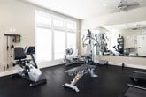 This fitness center is a thoughtfully designed, single-room facility that offers a comprehensive workout environment. The space features dark, durable flooring throughout, paired with neutral-toned walls and a textured ceiling equipped with recessed lighting and a ceiling fan to maintain comfort during exercise sessions.

Natural light floods the room through multiple large windows fitted with blinds, offering views of the surrounding outdoor environment. A full-length wall mirror spans a significant portion of one wall, effectively enhancing the sense of space and allowing users to monitor their form during workouts. A wall-mounted flat-screen television adds to the amenity offerings.

The fitness center is outfitted with a well-rounded selection of equipment. Cardio options include multiple treadmills, an elliptical machine, and a recumbent stationary bike. Strength training is supported by a multi-function cable and weight machine, a free-weight dumbbell rack, and a workout bench. Resistance bands are neatly organized on a wall-mounted rack, providing additional versatility for training routines.

Additional amenities include a shelving unit stocked with towels and supplies, a wall clock, and a direct-access exterior door leading to the outdoors. The layout is open and functional, allowing for ease of movement between equipment while maintaining a clean, organized atmosphere.