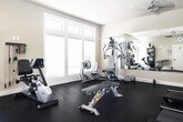 This fitness center is a thoughtfully designed, single-room facility that offers a comprehensive workout environment. The space features dark, durable flooring throughout, paired with neutral-toned walls and a textured ceiling equipped with recessed lighting and a ceiling fan to maintain comfort during exercise sessions.

Natural light floods the room through multiple large windows fitted with blinds, offering views of the surrounding outdoor environment. A full-length wall mirror spans a significant portion of one wall, effectively enhancing the sense of space and allowing users to monitor their form during workouts. A wall-mounted flat-screen television adds to the amenity offerings.

The fitness center is outfitted with a well-rounded selection of equipment. Cardio options include multiple treadmills, an elliptical machine, and a recumbent stationary bike. Strength training is supported by a multi-function cable and weight machine, a free-weight dumbbell rack, and a workout bench. Resistance bands are neatly organized on a wall-mounted rack, providing additional versatility for training routines.

Additional amenities include a shelving unit stocked with towels and supplies, a wall clock, and a direct-access exterior door leading to the outdoors. The layout is open and functional, allowing for ease of movement between equipment while maintaining a clean, organized atmosphere.