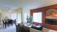 This single-floor space offers a welcoming and well-appointed interior designed for comfortable everyday living. The living area is the heart of the home, characterized by vaulted ceilings that lend an airy, open feel to the room. A striking terracotta-red accent wall adds a bold splash of color and visual depth, creating a warm and inviting atmosphere. Multiple windows dressed with vertical blinds allow an abundance of natural light to fill the space, brightening the neutral carpet flooring throughout. The living room is thoughtfully arranged with a dark leather sofa accented by earth-toned throw pillows, centered around a sleek black coffee table. A lush potted tropical plant adds a touch of greenery and life to the interior. Adjacent to the living area, a dining space is visible, furnished with dark wooden chairs and a table, providing a defined area for meals. The overall ambiance is casual yet polished, blending warm tones, natural elements, and practical furnishings into a cohesive and comfortable environment suitable for both relaxing and entertaining.