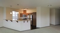 This single-floor residence offers a thoughtfully designed open-concept layout that seamlessly connects the kitchen and living areas. The kitchen is a standout feature, outfitted with stainless steel appliances including a refrigerator, range, and built-in microwave. Rich, warm-toned wood cabinetry extends to the ceiling, providing generous storage, while the peninsula countertop finished in granite features a breakfast bar overhang, making it ideal for casual dining or entertaining. A pendant light and a track lighting system illuminate the kitchen, enhancing both function and ambiance. The transition from the dark hardwood flooring in the kitchen to plush carpeting in the living area subtly defines each zone while maintaining an open, airy feel. A large window floods the space with natural light, contributing to the bright and welcoming atmosphere. The clean white walls and neutral palette throughout create a versatile backdrop that complements a variety of interior styles. Overall, the space balances modern finishes with practical design, making it a comfortable and functional single-level home.
