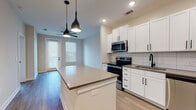 This single-story unit at Cadence Sugar Hill in Sugar Hill, Georgia, offers a well-designed open-concept layout that maximizes functionality. The kitchen serves as the centerpiece of the living area, featuring crisp white shaker cabinetry with matte black hardware, a white subway tile backsplash, gray countertops, and a full suite of stainless steel appliances including a range, microwave, dishwasher, and refrigerator. A central kitchen island with a light quartz surface provides additional prep space and casual seating, illuminated by two sleek pendant lights overhead. The kitchen flows seamlessly into the living area, which benefits from natural light through windows fitted with blinds and features wood-style flooring throughout. A ceiling fan enhances comfort in the main living space. The bedroom is carpeted and includes a window that allows natural light to fill the room. The bathroom is finished with wood-look flooring, a white vanity topped with granite countertops, matte black fixtures and hardware, a large framed mirror, and a tiled tub/shower combination. The overall design palette of neutral tones, modern hardware, and clean lines creates a cohesive and contemporary aesthetic throughout the unit.