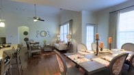 This thoughtfully designed two-story home offers a welcoming and well-appointed interior that blends comfort with classic elegance. The main level showcases an open-concept layout where the living and dining areas flow seamlessly together, creating a spacious and inviting atmosphere ideal for both everyday living and entertaining.

Rich hardwood flooring extends throughout the main living space, anchoring the warm, traditional aesthetic. The living area features elevated ceilings with a ceiling fan, adding to the sense of openness and airflow. Crown molding runs along the upper walls, lending a refined architectural detail to the space. Large windows dressed with white plantation shutters and rod-hung curtains allow generous natural light to fill the room while maintaining privacy.

The dining area accommodates a substantial dining table surrounded by upholstered chairs, positioned to take advantage of the natural light streaming through the windows. The open sightlines between the dining and living spaces enhance the overall sense of scale and connectivity.

The living room is furnished with classic seating arrangements, complemented by layered area rugs and tasteful decorative accents that create a cozy yet polished ambiance. The second floor adds additional living space, completing this well-rounded and functional home.