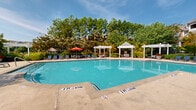 Located at 10840 State Bridge Rd in Alpharetta, Georgia, this outdoor amenity area centers around a generously sized swimming pool with crystal-clear turquoise water. The pool deck is finished in a smooth concrete surround and is equipped with multiple blue lounge chairs and tables, offering ample space for relaxation. Vibrant red umbrellas provide shaded seating areas, adding a welcoming pop of color throughout the space. Two elegant white pergolas serve as covered gathering spots, framing the poolside experience and providing additional shade and structure. The area is beautifully landscaped, with mature trees, flowering shrubs, and seasonal plantings creating a lush, park-like backdrop that enhances the sense of privacy and natural serenity. A charming brick clubhouse building with a pitched roof is visible adjacent to the pool, complementing the residential architectural character of the community. The pool entry features clearly marked depth indicators and safety signage, reflecting a well-maintained and safety-conscious facility. Surrounded by tall Georgia pines and hardwoods, the space enjoys an open-air ambiance that feels both expansive and sheltered, making it an inviting outdoor retreat for all seasons.
