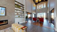 This thoughtfully designed single-floor amenity space offers an impressive blend of modern aesthetics and functional comfort. The centerpiece of the space is a dramatic great room with soaring double-height ceilings, eye-catching pendant lighting in varied shapes and finishes, and a combination of large-format tile and warm hardwood flooring. A feature wall clad in a textured geometric wallcovering anchors a built-in linear fireplace and wall-mounted television, flanked by floor-to-ceiling open shelving displaying curated décor. The lounge seating area includes bold red armchairs and a plush upholstered bench, creating an inviting atmosphere for gathering and relaxation.

Adjacent to the lounge, a café-style bar and kitchen area showcases a striking iridescent teal tile accent wall, dark cabinetry, and a substantial marble-look island with bar seating — ideal for socializing or casual dining. The dining area, framed by expansive black-framed windows and glass doors, offers panoramic views of an outdoor pool and courtyard, flooding the room with natural light.

Compact collaboration nooks with backlit wood-panel privacy screens, white-topped tables, and mustard-yellow ottomans provide quiet, focused spaces. Throughout, curated artwork, layered textures, and a cohesive warm-neutral palette contribute to a sophisticated yet welcoming communal environment.