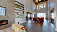 This thoughtfully designed single-floor amenity space offers an impressive blend of modern aesthetics and functional comfort. The centerpiece of the space is a dramatic great room with soaring double-height ceilings, eye-catching pendant lighting in varied shapes and finishes, and a combination of large-format tile and warm hardwood flooring. A feature wall clad in a textured geometric wallcovering anchors a built-in linear fireplace and wall-mounted television, flanked by floor-to-ceiling open shelving displaying curated décor. The lounge seating area includes bold red armchairs and a plush upholstered bench, creating an inviting atmosphere for gathering and relaxation.

Adjacent to the lounge, a café-style bar and kitchen area showcases a striking iridescent teal tile accent wall, dark cabinetry, and a substantial marble-look island with bar seating — ideal for socializing or casual dining. The dining area, framed by expansive black-framed windows and glass doors, offers panoramic views of an outdoor pool and courtyard, flooding the room with natural light.

Compact collaboration nooks with backlit wood-panel privacy screens, white-topped tables, and mustard-yellow ottomans provide quiet, focused spaces. Throughout, curated artwork, layered textures, and a cohesive warm-neutral palette contribute to a sophisticated yet welcoming communal environment.