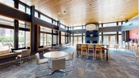 Located at 101 Summit St in Duluth, MN, this single-floor community lounge is a thoughtfully designed gathering space that blends contemporary aesthetics with functional comfort. The room is defined by its dramatic double-height ceilings finished in warm wood paneling, which contrasts beautifully with the dark-framed floor-to-ceiling windows that wrap around the perimeter, flooding the interior with natural light and providing views of the surrounding outdoor areas.

A striking stacked-stone fireplace serves as the room's focal point, paired with a wall-mounted television and a large sectional sofa, creating an inviting lounge area ideal for relaxation or social gatherings. Stylish pendant lighting complements the warm tones of the wood ceiling throughout. Built-in window seat nooks with cushioned benches offer additional casual seating tucked beneath the expansive glazing.

The space accommodates multiple functions, featuring round café-style tables with modern chairs and a tall communal bar-height table for collaborative or dining use. Patterned carpet tiles in cool blue-gray tones ground the open floor plan. A private glass-enclosed conference room offers a dedicated workspace, while a small kitchenette with a sink and beverage station provides added convenience. Together, these elements make this space a versatile, well-appointed community amenity.