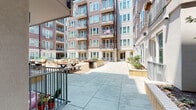 This ground-floor space is situated within a contemporary multi-story residential building, offering a unique blend of private outdoor living and access to vibrant shared community amenities. The unit opens directly onto a private patio through a sliding glass door, providing a seamless indoor-outdoor connection. Adjacent to the patio, a thoughtfully landscaped garden area features ornamental trees, shrubs, and decorative rock borders set against a mulched bed, creating a tranquil natural retreat.

The exterior of the building showcases a modern architectural design with horizontal wood-toned cladding, teal-framed windows, and individual balconies on the upper floors. The building's interior courtyard, accessible from the unit, is a spacious open-air communal area finished with large-format concrete pavers. It is well-appointed with picnic tables, lounge seating arrangements, raised planter beds, and mature landscaping, making it an inviting gathering space for residents.

Smooth stucco exterior walls in warm neutral tones frame the private patio, which is finished with clean tile flooring. The overall ambiance is bright and airy, benefiting from abundant natural sunlight throughout the day. The combination of private garden space, direct courtyard access, and modern architectural detailing makes this a distinctive ground-level offering within its residential community.
