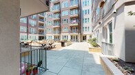 This ground-floor space is situated within a contemporary multi-story residential building, offering a unique blend of private outdoor living and access to vibrant shared community amenities. The unit opens directly onto a private patio through a sliding glass door, providing a seamless indoor-outdoor connection. Adjacent to the patio, a thoughtfully landscaped garden area features ornamental trees, shrubs, and decorative rock borders set against a mulched bed, creating a tranquil natural retreat.

The exterior of the building showcases a modern architectural design with horizontal wood-toned cladding, teal-framed windows, and individual balconies on the upper floors. The building's interior courtyard, accessible from the unit, is a spacious open-air communal area finished with large-format concrete pavers. It is well-appointed with picnic tables, lounge seating arrangements, raised planter beds, and mature landscaping, making it an inviting gathering space for residents.

Smooth stucco exterior walls in warm neutral tones frame the private patio, which is finished with clean tile flooring. The overall ambiance is bright and airy, benefiting from abundant natural sunlight throughout the day. The combination of private garden space, direct courtyard access, and modern architectural detailing makes this a distinctive ground-level offering within its residential community.