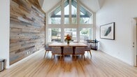 This thoughtfully designed single-floor space blends mid-century modern aesthetics with warm, rustic elements to create an inviting and functional environment. The main living area commands attention with its soaring vaulted ceilings and an expansive wall of floor-to-ceiling windows and clerestory glazing that floods the room with natural light. A tile-surround fireplace serves as the focal point, flanked by sculptural lounge chairs and anchored by a bold blue area rug, while a mounted flat-screen TV and French doors leading to an outdoor deck complete the space.

The open dining area features a substantial wood dining table surrounded by upholstered chairs, set beneath the dramatic gabled ceiling. A full-height reclaimed wood accent wall adds rich texture, and the large windows frame views of the outdoor deck and surrounding greenery.

The kitchen is well-appointed with dark cabinetry, stainless steel appliances including a full-size refrigerator, oven, microwave, and dishwasher, complemented by a striking patterned tile backsplash and pendant lighting.

A cozy lounge nook, separated by distinctive teal sliding barn doors, features reclaimed wood wall and ceiling cladding, built-in bench seating, round bistro tables, and teal accent chairs — ideal for intimate gatherings. Light hardwood flooring flows seamlessly throughout, unifying the entire space.