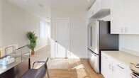 This single-floor space offers a clean, modern aesthetic characterized by crisp white walls, light hardwood flooring, and an abundance of natural light streaming through large windows fitted with horizontal blinds. The open layout seamlessly connects the kitchen and main living or work area, creating a versatile and functional environment well-suited for a live-work setup.

The kitchen is well-appointed with stainless steel appliances, including a French door refrigerator, a gas range, and an over-the-range microwave. White shaker-style cabinetry extends to the ceiling, maximizing storage, while light-toned countertops contribute to the space's bright and airy feel. The adjacent main area accommodates a dedicated workspace with room for a desk, chairs, and office equipment, all bathed in natural light from multiple windows. A glass-paneled door provides direct access to the exterior, enhancing the sense of openness.

The bathroom is finished in a neutral palette, featuring light hardwood flooring consistent with the rest of the space, a white vanity with a stone-look countertop, a large mirror, and a glass-enclosed shower. Recessed lighting throughout the space adds to the clean, contemporary character. Overall, the space strikes a balance between practicality and modern design, offering a bright and welcoming atmosphere suited for a range of uses.