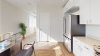 This single-floor space offers a clean, modern aesthetic characterized by crisp white walls, light hardwood flooring, and an abundance of natural light streaming through large windows fitted with horizontal blinds. The open layout seamlessly connects the kitchen and main living or work area, creating a versatile and functional environment well-suited for a live-work setup.

The kitchen is well-appointed with stainless steel appliances, including a French door refrigerator, a gas range, and an over-the-range microwave. White shaker-style cabinetry extends to the ceiling, maximizing storage, while light-toned countertops contribute to the space's bright and airy feel. The adjacent main area accommodates a dedicated workspace with room for a desk, chairs, and office equipment, all bathed in natural light from multiple windows. A glass-paneled door provides direct access to the exterior, enhancing the sense of openness.

The bathroom is finished in a neutral palette, featuring light hardwood flooring consistent with the rest of the space, a white vanity with a stone-look countertop, a large mirror, and a glass-enclosed shower. Recessed lighting throughout the space adds to the clean, contemporary character. Overall, the space strikes a balance between practicality and modern design, offering a bright and welcoming atmosphere suited for a range of uses.