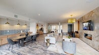 This single-story clubhouse in Gonzales, Louisiana offers a well-appointed, open-concept interior designed to serve as a premier community gathering space. Wood-look tile flooring flows throughout, unifying the various functional zones while lending a warm, contemporary aesthetic.

The main lounge area is anchored by a floor-to-ceiling marble-clad fireplace wall, which also accommodates a wall-mounted television. Plush seating arrangements, including curved accent chairs and a neutral sofa, are complemented by sculptural side tables and a statement gold chandelier overhead. A built-in banquette with a slatted wood accent wall provides an intimate nook with seating, positioned alongside large glass doors that invite natural light and outdoor views.

The dining area features a substantial dark wood communal table paired with modern side chairs, positioned adjacent to a fully equipped kitchen. The kitchen showcases warm wood cabinetry, a two-tone subway tile backsplash in white and soft blue, stainless steel appliances, and a row of brass pendant lights that add a refined touch. A small built-in workstation alcove with a bold red pendant light offers a practical spot for quick tasks. The entry corridor leads to additional rooms, completing this versatile and stylishly designed clubhouse.