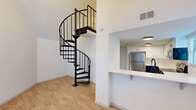 Located in the heart of downtown Austin at 205 E 4th St, this two-story residence offers a thoughtfully designed layout across two floors. The main level serves as the social hub of the home, featuring a spacious living area with soaring ceilings and warm wood-look flooring throughout. A standout architectural element is the striking wrought-iron spiral staircase, which rises elegantly to the upper level and adds a bold visual focal point to the open living space. The kitchen is designed for both function and connection, featuring white cabinetry, stainless steel appliances including a full-size refrigerator, and a pass-through counter with a breakfast bar that opens directly to the living area, allowing natural light from nearby windows to flow through. The upper level hosts the generously sized primary bedroom, which features a ceiling fan with integrated lighting, crown molding, a window with natural light, and direct access to the full bathroom. The bathroom is finished with matte black fixtures, including a tub/shower combination and a stylish vanity with a round mirror and navy blue cabinetry. Clean white walls and consistent flooring throughout create a cohesive, modern aesthetic ideal for urban living.