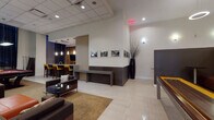 This single-floor amenity space is thoughtfully designed to accommodate a variety of social activities across several distinct zones. The main lounge area is anchored by a striking marble-finished feature wall with a wall-mounted TV flanked by lush potted plants and warm wood cabinetry. Two large, low-profile orange sofas face each other across a minimalist coffee table, creating an inviting gathering space with floor-to-ceiling windows that bring in natural views. An adjacent lounge area features dark leather seating, bold accent pillows in mustard and grey tones, wall sconces, and black-and-white urban photography on deep charcoal walls, lending a more intimate atmosphere. A raised bar and kitchen area includes a granite-topped island with yellow upholstered stools, pendant lighting, and a distinctive wine crate accent wall with a mounted flat-screen TV. The games area accommodates a regulation-style billiards table alongside comfortable lounge seating and a TV nook with warm wood paneling. Throughout the space, light tile flooring, recessed ceiling lighting, neutral area rugs, and decorative architectural details maintain a cohesive, upscale contemporary aesthetic designed for communal enjoyment.