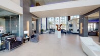 This thoughtfully designed amenity space makes a striking first impression with its grand, double-height lobby. Soaring floor-to-ceiling glass walls flood the interior with natural light while offering views of the surrounding exterior. A sculptural bronze art piece commands the center of the lobby, complemented by warm wood slat accent walls, large-format tile flooring, and a carefully curated seating arrangement that balances elegance with comfort. A dramatic nature-inspired mural featuring tropical trees and vibrant parrots adds an artistic focal point to one of the lounge areas, which is furnished with plush sofas and contemporary accent chairs. Adjacent spaces include a sleek bar and kitchen area with a gold-tiled island, pendant lighting, and views toward an outdoor pool area, as well as a conference or dining room outfitted with a long table, modern pendant fixtures, and a mosaic tile media wall. The fitness center is fully equipped with a comprehensive array of cardio and strength machines, accented by a dark ceiling, circular pendant lighting, and large windows overlooking palm trees. An elevator lobby with a textured dark feature wall, linear ceiling lighting, and designer seating provides refined circulation throughout the building. Each area reflects a cohesive modern aesthetic with high-end finishes throughout.