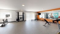 Located at 1515 Penn Ave in Wilkinsburg, PA, this fitness center offers a well-equipped exercise space designed to support a comprehensive workout routine. The room is finished with durable rubber flooring throughout, providing a safe and practical surface ideal for gym use.

The space is distinctively styled with bold accent walls in teal and orange, creating an energetic and motivating atmosphere. Recessed flush lighting keeps the room bright and evenly illuminated, while large sliding glass doors along one wall invite natural light and offer views of the surrounding outdoor greenery.

The exercise room is outfitted with a diverse selection of Matrix-brand equipment, including a treadmill, rowing machine, cable resistance machine, and elliptical trainer. Spinning bikes with digital displays provide dedicated cardio options, while a full dumbbell rack with tiered weight storage supports strength training. A weight bench and stability balls round out the available equipment.

A large wall-mounted mirror on the orange accent wall enhances the sense of space and allows users to monitor their form during workouts. Decorative artwork adds a personal touch to the teal feature wall. The layout is open and functional, allowing multiple users to exercise simultaneously with ease. A flat-screen TV mounted on the wall adds a convenient entertainment option during workouts.