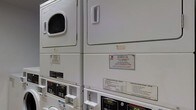 This single-floor laundry facility is equipped with commercial-grade, coin-operated stacked washer and dryer units, serviced and maintained by Coinmach. The machines are arranged in a row, maximizing the use of vertical space with a stacked configuration that accommodates multiple units within a compact footprint. Each unit features clearly labeled operating instructions for both the washer and dryer, ensuring straightforward use. The dryers display digital readouts, and the machines accept coin payments, as indicated by the coin slots and pricing displays visible on the control panels. Service contact information is prominently posted on each unit, directing users to Coinmach's customer support line and website for any maintenance concerns. The facility is designed for practical, efficient use, providing residents or users with a reliable and accessible laundry solution on-site. The overall layout and equipment suggest a well-organized, functional shared laundry space suited for a residential building or similar multi-unit property.