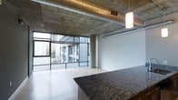 This single-floor space showcases a striking industrial-modern aesthetic that blends raw architectural elements with contemporary finishes. The exposed concrete ceiling is a defining feature, complemented by visible ductwork and ventilation systems that add to the loft-like character of the space. A pendant light fixture hangs from the ceiling, providing focused illumination and a subtle design accent.

The open-plan layout is anchored by a substantial granite countertop in a rich, dark tone, which serves as both a functional and visual centerpiece. The light hardwood flooring contrasts effectively with the raw concrete overhead, creating a balanced interplay of warm and industrial materials.

Floor-to-ceiling windows with a grid-frame design span a generous portion of the perimeter, flooding the interior with natural light while offering expansive views of the surrounding urban landscape, including neighboring buildings and city greenery. A structural concrete column is thoughtfully integrated into the layout without disrupting the open feel of the space.

White walls provide a clean, neutral backdrop that allows the architectural features to take center stage, resulting in an overall ambiance that is modern, urban, and visually dynamic.