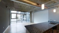This single-floor space showcases a striking industrial-modern aesthetic that blends raw architectural elements with contemporary finishes. The exposed concrete ceiling is a defining feature, complemented by visible ductwork and ventilation systems that add to the loft-like character of the space. A pendant light fixture hangs from the ceiling, providing focused illumination and a subtle design accent.

The open-plan layout is anchored by a substantial granite countertop in a rich, dark tone, which serves as both a functional and visual centerpiece. The light hardwood flooring contrasts effectively with the raw concrete overhead, creating a balanced interplay of warm and industrial materials.

Floor-to-ceiling windows with a grid-frame design span a generous portion of the perimeter, flooding the interior with natural light while offering expansive views of the surrounding urban landscape, including neighboring buildings and city greenery. A structural concrete column is thoughtfully integrated into the layout without disrupting the open feel of the space.

White walls provide a clean, neutral backdrop that allows the architectural features to take center stage, resulting in an overall ambiance that is modern, urban, and visually dynamic.