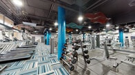 Located at 5225 Blakeslee Ave in Los Angeles, CA, this fitness center offers a comprehensive and thoughtfully designed workout environment across a single, expansive floor. The space is characterized by an industrial-chic aesthetic, with exposed ductwork and dark ceilings that contrast sharply against vibrant blue structural columns and playful, multicolored wave-shaped ceiling sculptures that add a dynamic artistic element to the interior.

The flooring transitions strategically between zones: a bold, patterned blue-and-white carpet defines the cardio and stretching areas, while durable rubber flooring covers the free weights and strength training sections. Large floor-to-ceiling windows along one wall flood the space with natural light and offer views of the surrounding streetscape, creating an open, energizing atmosphere.

The cardio zone is well-appointed with a row of treadmills, elliptical trainers, and stationary cycling bikes, many equipped with personal display screens. The strength training area features an extensive dumbbell rack, multiple cable machines, and a variety of selectorized equipment. Full-length mirrors line key walls, enhancing the sense of space and allowing for proper form checks during workouts. Recessed and track lighting ensure the entire floor is brightly and evenly illuminated, supporting a focused and motivating fitness experience.