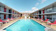 This single-story property is centered around a spacious outdoor pool area framed by a U-shaped building with a classic red-and-white exterior. The courtyard-style pool deck is outfitted with red lounge chairs and potted tropical plants, creating a relaxed, resort-like atmosphere open to the sky. The surrounding structure features covered walkways and balcony-style railings, giving the outdoor common area a welcoming, enclosed feel.

The fitness center is a standout amenity, spread across a generously sized space with soaring ceilings and bright white walls that create an open, airy environment. Durable rubber-speckled flooring runs throughout, providing a functional surface suited to heavy use. The facility is divided into distinct workout zones: one area houses cardio equipment, including treadmills, stationary bikes, and an elliptical, all positioned beneath wall-mounted televisions. A separate strength-training section features Life Fitness cable machines, a lat pulldown station, free weights, adjustable benches, and a tire sled. Floor-to-ceiling mirrors along one wall expand the space visually. French doors with glass panels provide natural light at the entrance, reinforcing the clean, professional character of the facility.