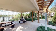 This single-level outdoor space centers around a generously sized covered patio area designed for communal gathering and relaxation. The structure is defined by a clean gabled roof supported by warm-toned square columns, providing ample shade over a smooth concrete floor. A round picnic table with attached bench seating sits beneath the overhang, offering a casual dining or social setting. Adjacent to the main covered area, a decorative pergola with thatched accents adds a tropical, resort-like character to the space. The patio is bordered by a black metal railing, beyond which a richly landscaped greenway unfolds, featuring mature palm trees, dense shrubs, and vibrant foliage that lend the area a lush, verdant backdrop. The surrounding vegetation creates a sense of privacy and natural enclosure while allowing open sightlines to the greenery beyond. The overall ambiance is relaxed and inviting, evoking a warm, Southern California or tropical aesthetic that is well-suited for outdoor leisure. Additional amenities visible near the perimeter suggest the space is equipped for outdoor use and convenience. The combination of structured shelter and open-air surroundings makes this a versatile and appealing communal outdoor environment.