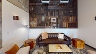 Located at 4040 26th Ave SW in Seattle, WA, this single-floor commercial space masterfully blends industrial design with warm, inviting aesthetics. The open-plan layout is anchored by a striking double-sided gas fireplace clad in large-format metallic tiles, topped with a sculptural art piece that serves as a dramatic focal point between zones.

Exposed concrete ceilings with visible ductwork reinforce the industrial character throughout, while polished concrete floors provide a sleek, durable foundation. A show-stopping reclaimed wood accent wall — composed of varied timber panels, hardware, and textures — adds artistic depth to the lounge area, which is furnished with a tufted leather sofa and natural wood side tables.

The dining and collaborative area features a live-edge communal table, surrounded by mixed seating and illuminated by an iconic Sputnik-style chandelier. Mustard yellow booth seating lines one wall, complemented by Edison bulb pendant lighting for a warm, inviting ambiance. An exposed brick wall with steel-framed clerestory windows allows natural light to filter in from the exterior greenery beyond.

A glass-partitioned private office or meeting room provides separation without sacrificing visual openness. Industrial-style shelving units with rolling ladders offer both storage and display. The overall atmosphere is sophisticated yet approachable — a thoughtfully curated workspace designed for productivity and collaboration.